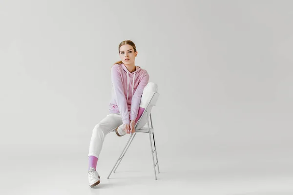 Schöne junge Frau in rosa Kapuzenpulli sitzt auf Stuhl auf weiß und blickt in die Kamera — Stockfoto