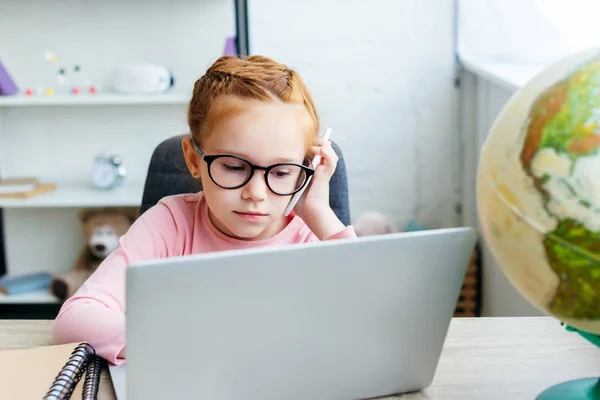 Сфокусированный ребенок в очках разговаривает со смартфоном и использует ноутбук дома — стоковое фото