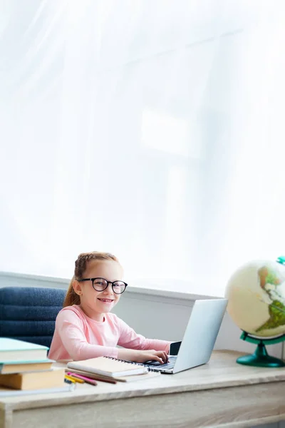 Adorable niño en gafas de vista sonriendo a la cámara mientras estudia con el ordenador portátil en casa - foto de stock