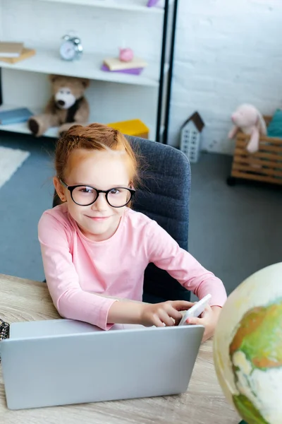 High angle view of adorable red haired schoolchild in eyeglasses smiling at camera while using smartphone and laptop — Stock Photo