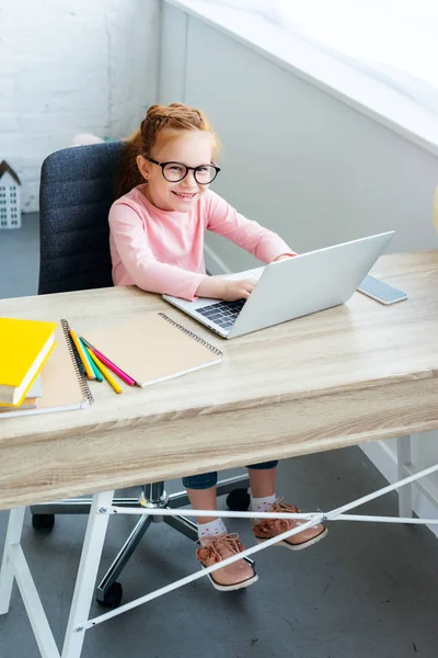 High angle view of beautiful kid in eyeglasses using laptop and smiling at camera — Stock Photo