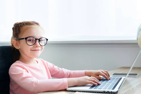 Hermoso niño en gafas con el ordenador portátil y sonriendo a la cámara - foto de stock
