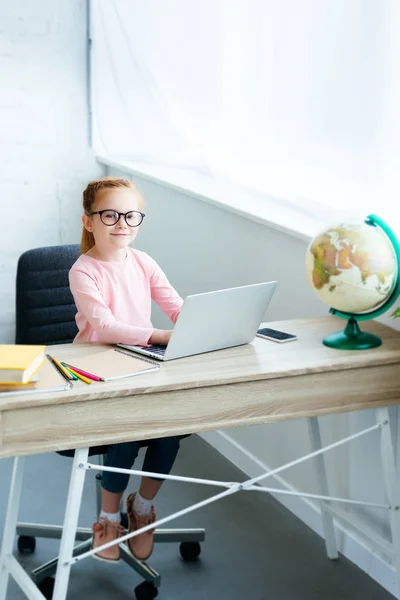 Vue grand angle de l'enfant dans les lunettes souriant à la caméra tout en utilisant un ordinateur portable et étudier à la maison — Photo de stock