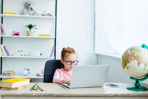 Cute little schoolchild in eyeglasses using laptop while studying at home — Stock Photo