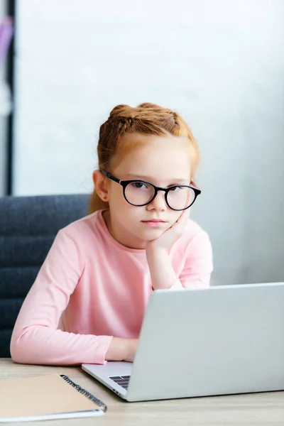 Bored little schoolchild in eyeglasses looking at camera while srtudying with laptop — Stock Photo