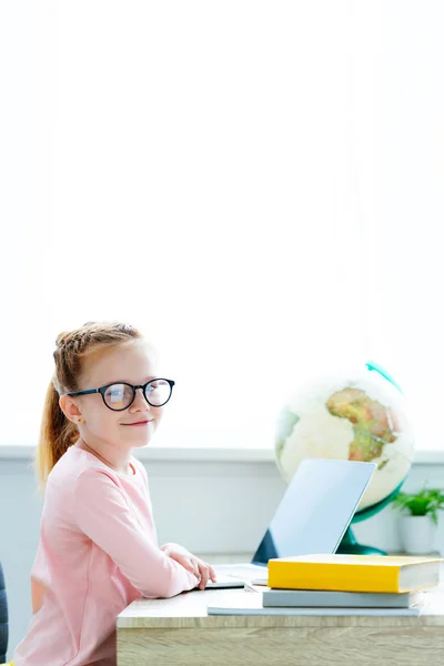 Hermosa colegiala pelirroja en gafas sonriendo a la cámara mientras estudia con libros y portátil en el escritorio - foto de stock