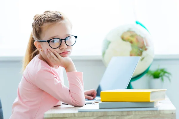 Скучающий ребенок в очках, смотрящий в камеру во время учебы с ноутбуком и книгами — стоковое фото