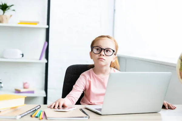 Thoughtful little schoolchild in eyeglasses looking up while sitting at desk and using laptop — Stock Photo