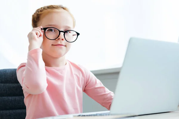 Красива шкільна дитина регулює окуляри і посміхається на камеру при використанні ноутбука — стокове фото