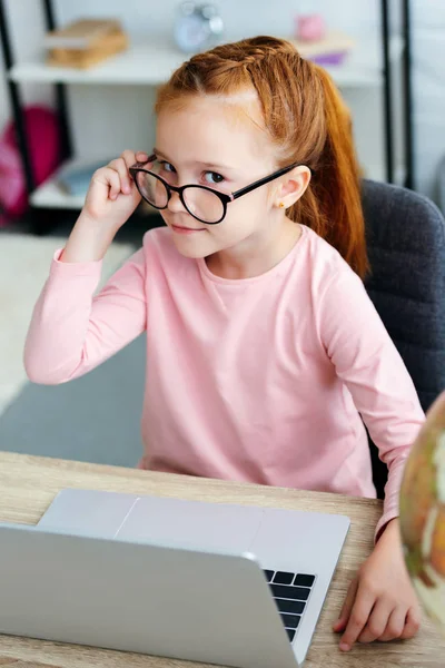 Vista de ángulo alto de hermosa colegiala ajustando las gafas y sonriendo a la cámara mientras usa el ordenador portátil - foto de stock