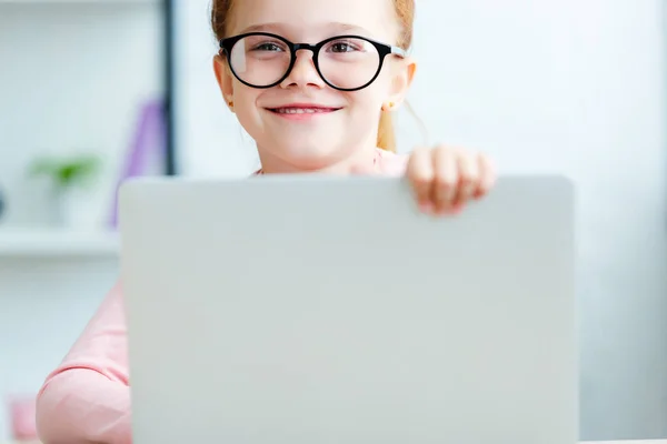 Adorable red haired schoolgirl in eyeglasses using laptop and smiling at camera — Stock Photo