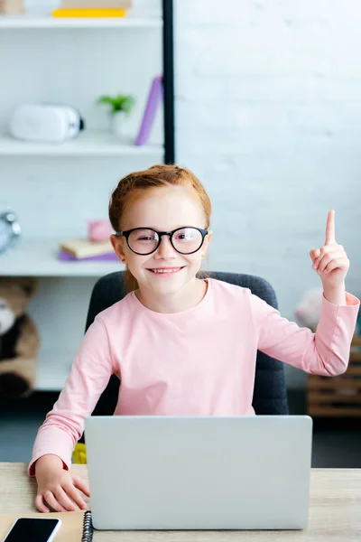 Adorable colegiala pelirroja en gafas apuntando hacia arriba con el dedo y sonriendo a la cámara mientras usa el ordenador portátil en casa - foto de stock