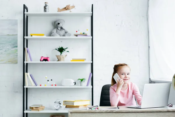 Belle rousse enfant parler par smartphone et sourire à la caméra tout en utilisant un ordinateur portable — Photo de stock