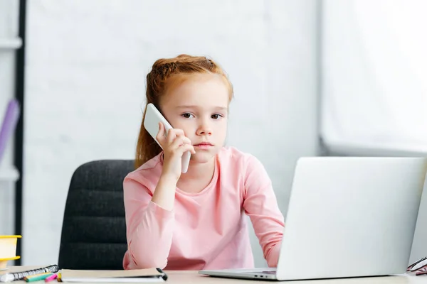 Belle rousse enfant parler par smartphone et regarder la caméra tout en utilisant un ordinateur portable — Photo de stock