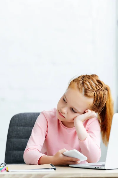 Niño pelirrojo aburrido mirando hacia otro lado mientras se utiliza el teléfono inteligente y portátil - foto de stock