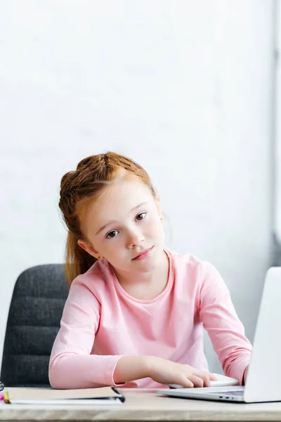 Gelangweilte kleine Schülerin schaut auf Kamera, während sie zu Hause Laptop benutzt — Stockfoto