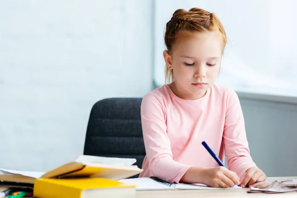 Adorable red haired schoolgirl sitting at desk and studying at home — Stock Photo