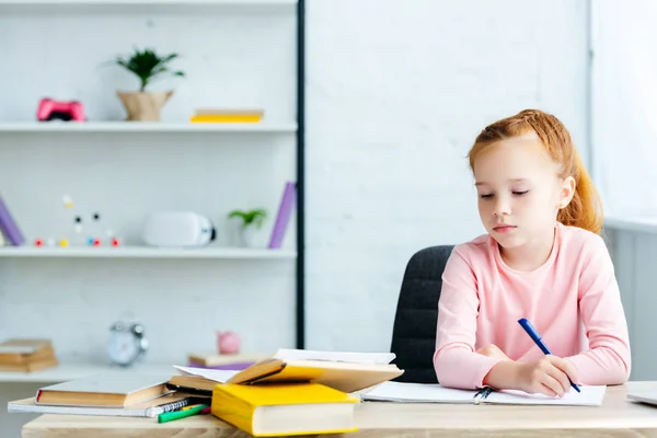 Belle écolière rousse assise au bureau et étudiant à la maison — Photo de stock