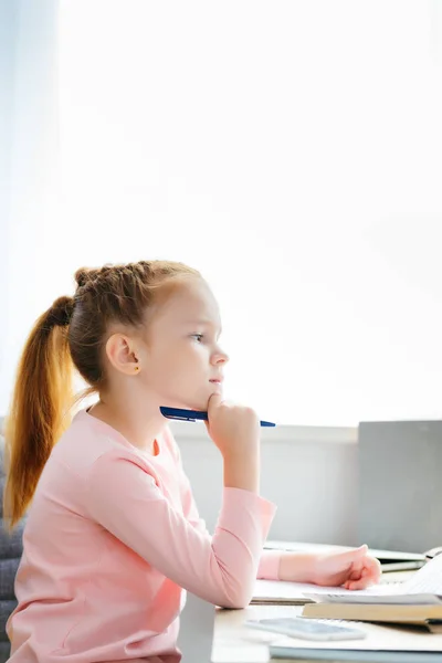 Vista laterale della studentessa premurosa che tiene la penna e distoglie lo sguardo mentre studia a casa — Foto stock