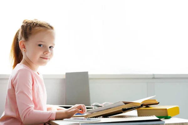 Hermosa colegiala sonriendo a la cámara mientras estudia en casa - foto de stock