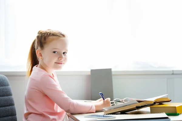 Seitenansicht der schönen Schülerin, die in die Kamera lächelt, während sie am Schreibtisch in Notizbuch schreibt — Stockfoto