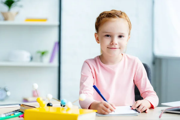 Schöne rothaarige Schülerin lächelt in die Kamera, während sie am Schreibtisch in ein Notizbuch schreibt — Stockfoto