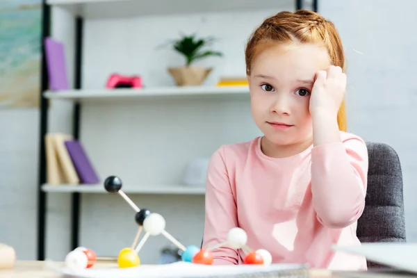 Hermosa colegiala pelirroja mirando a la cámara mientras estudia con modelo molecular en casa - foto de stock