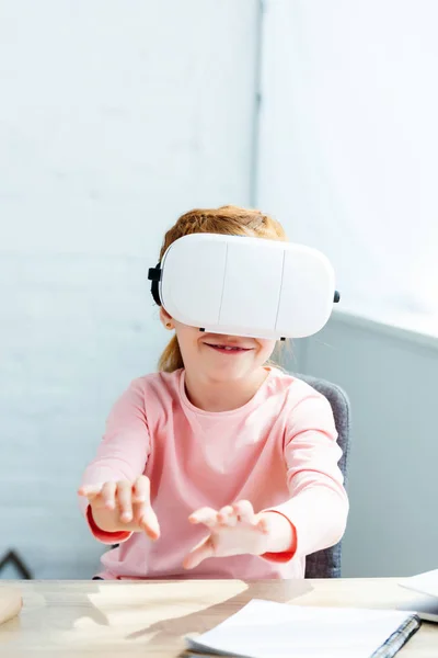 Sorrindo pequena criança em idade escolar usando fone de ouvido realidade virtual enquanto estudava em casa — Fotografia de Stock