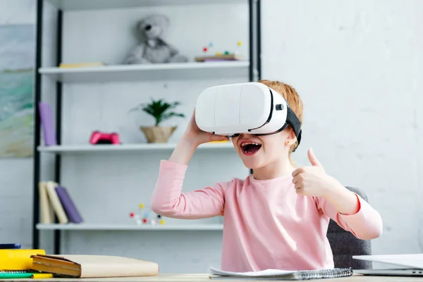 Adorable feliz escolar usando auriculares de realidad virtual y mostrando el pulgar hacia arriba - foto de stock