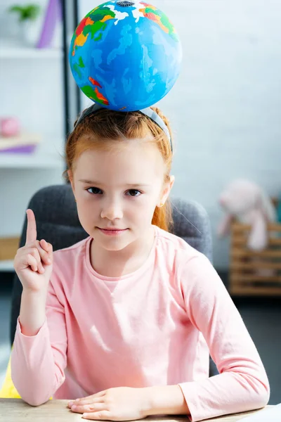Belle écolière avec globe sur la tête regardant la caméra et pointant vers le haut avec le doigt — Photo de stock