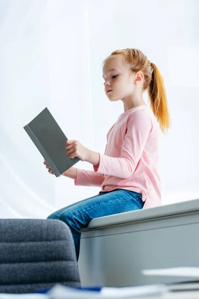 Низкий угол зрения симпатичной маленькой школьницы, сидящей на подоконнике и читающей книгу — стоковое фото