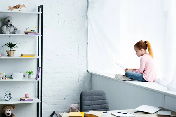 Beautiful redhead schoolgirl using digital tablet while sitting on windowsill — Stock Photo