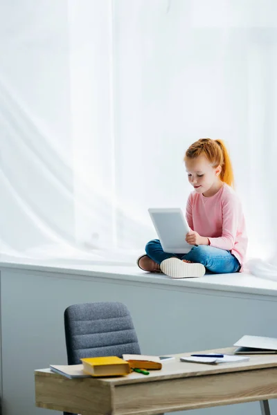 Adorable child using digital tablet while sitting on windowsill — Stock Photo