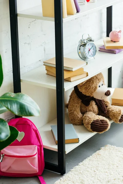 Livros, despertador e ursinho de pelúcia nas prateleiras e saco escolar rosa no tapete — Fotografia de Stock