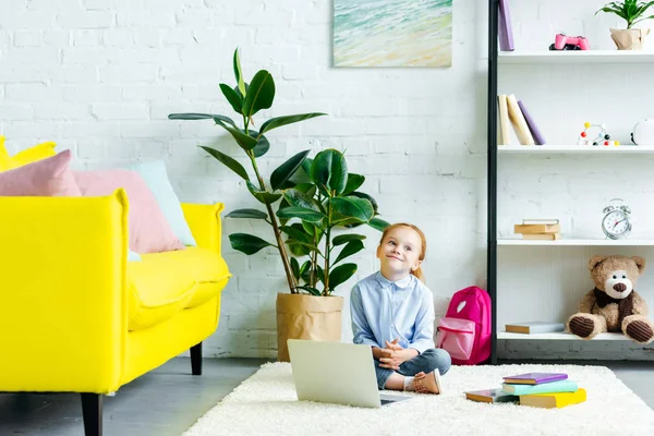 Petite écolière souriante regardant loin tout en étudiant avec ordinateur portable et livres à la maison — Photo de stock