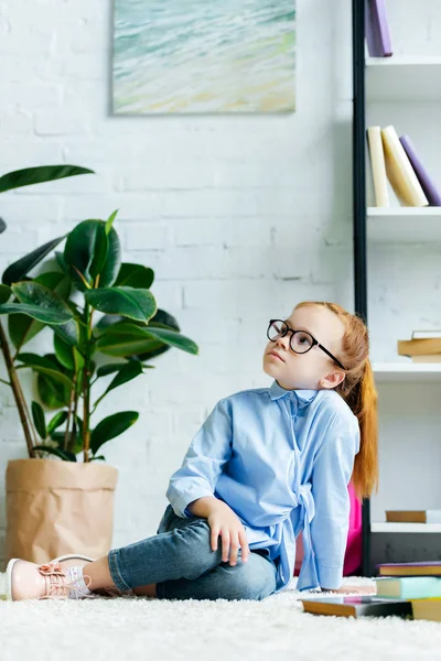 Belle petite rousse enfant en lunettes assis sur le tapis et levant les yeux tout en étudiant à la maison — Photo de stock