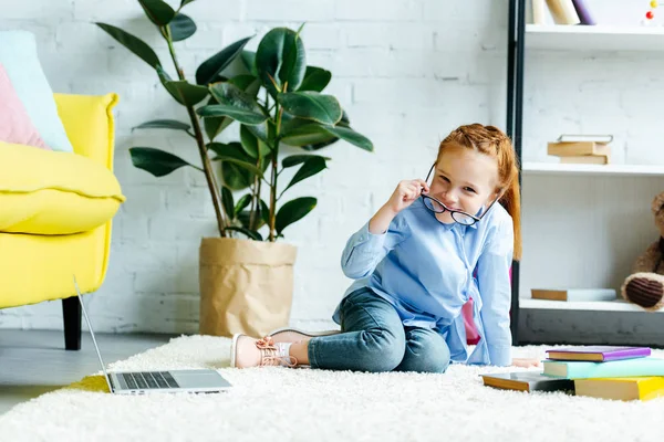 Bambino felice con gli occhiali che sorride alla macchina fotografica mentre studia con libri e laptop a casa — Foto stock