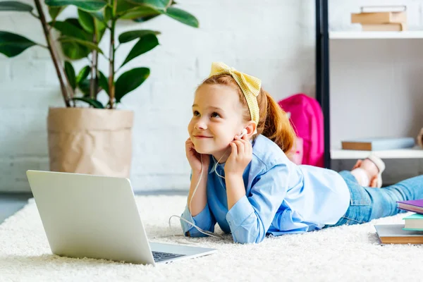 Cher enfant souriant dans les écouteurs levant les yeux tout en étant couché sur le tapis et en utilisant un ordinateur portable à la maison — Photo de stock