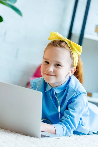 Hermoso niño de pelo rojo en auriculares sonriendo a la cámara mientras está acostado en la alfombra y el uso de la computadora portátil - foto de stock