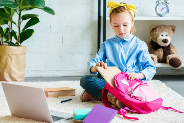 Cute little child packing school bag while sitting on carpet at home — Stock Photo