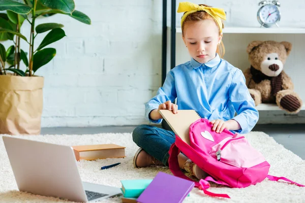 Mignon petit enfant emballant sac d'école tout en étant assis sur le tapis à la maison — Photo de stock