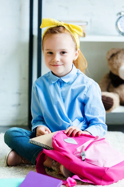 Entzückendes Kind lächelt in die Kamera, während es auf dem Teppich sitzt und Schultasche packt — Stockfoto