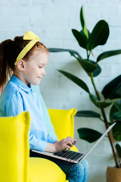 Vista laterale del bambino rosso sorridente seduto e utilizzando il computer portatile — Foto stock