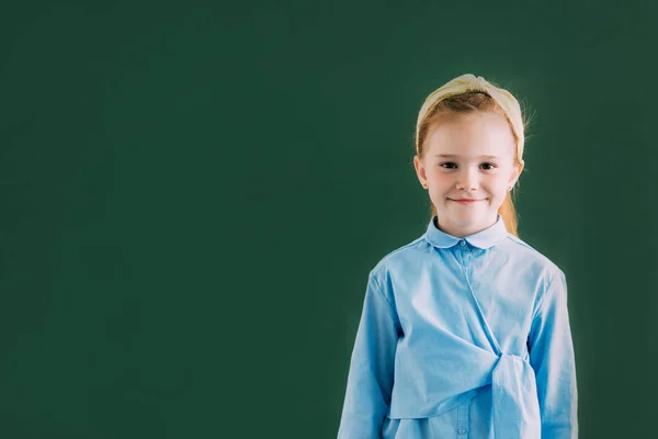Adorable little redhead schoolgirl standing near blackboard and smiling at camera — Stock Photo