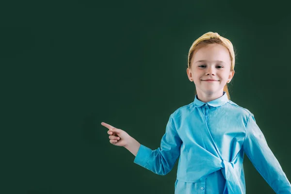 Beautiful red haired schoolgirl pointing at blank chalkboard and smiling at camera — Stock Photo