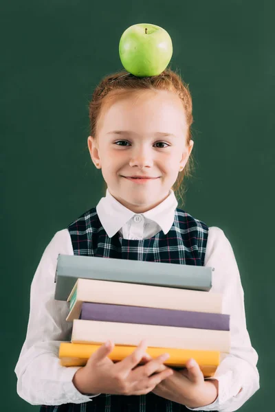 Adorable little redhead schoolgirl with apple on head holding pile of books and smiling at camera — Stock Photo