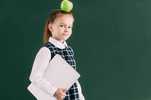 Красивая маленькая школьница с яблоком на голове держит ноутбук и смотрит в камеру — стоковое фото