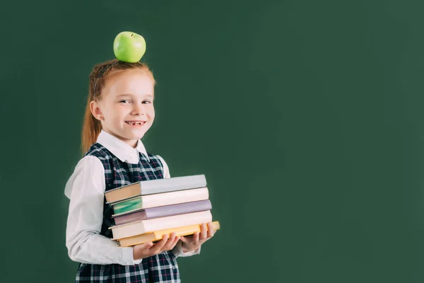 Beautiful little schoolgirl with apple on head holding pile of books and smiling at camera — Stock Photo