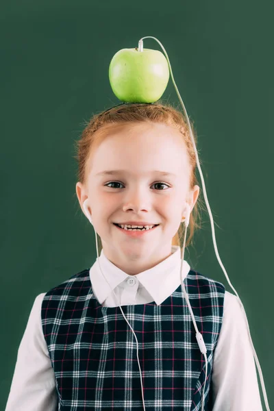 Pequena estudante feliz com maçã na cabeça e fones de ouvido sorrindo e olhando para a câmera — Fotografia de Stock