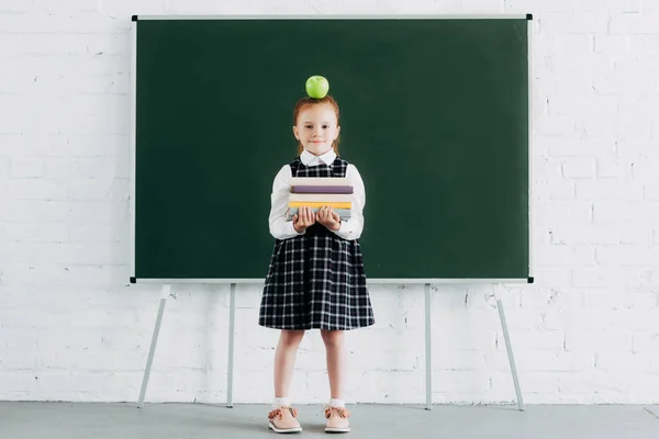 Nettes kleines Schulmädchen mit Apfel auf dem Kopf, das einen Stapel Bücher hält und in die Kamera schaut, während es neben der Tafel steht — Stockfoto
